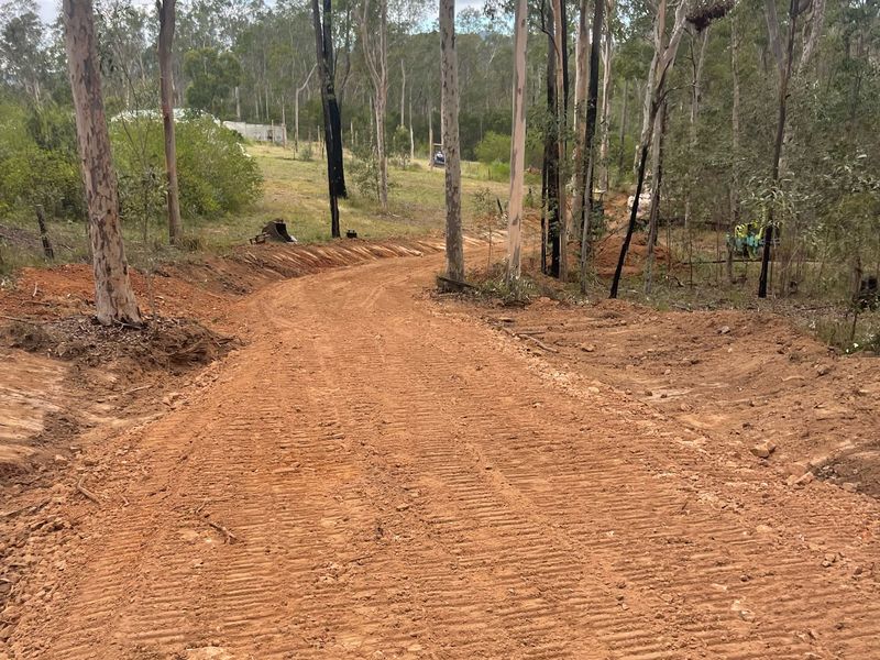 The same rural driveway after cutting, shaping and drainage: a wider red-earth surface with machine track marks